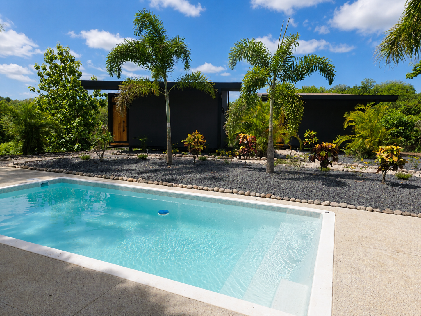 Swimming pool with clear turquoise water, tropical palms and the two dark-sand bungalows in the background