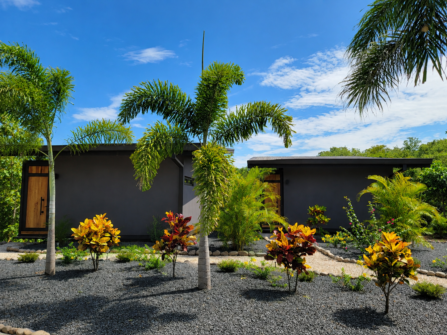 Two dark-sand bungalows set among tropical palms and landscaped gardens under a bright blue sky